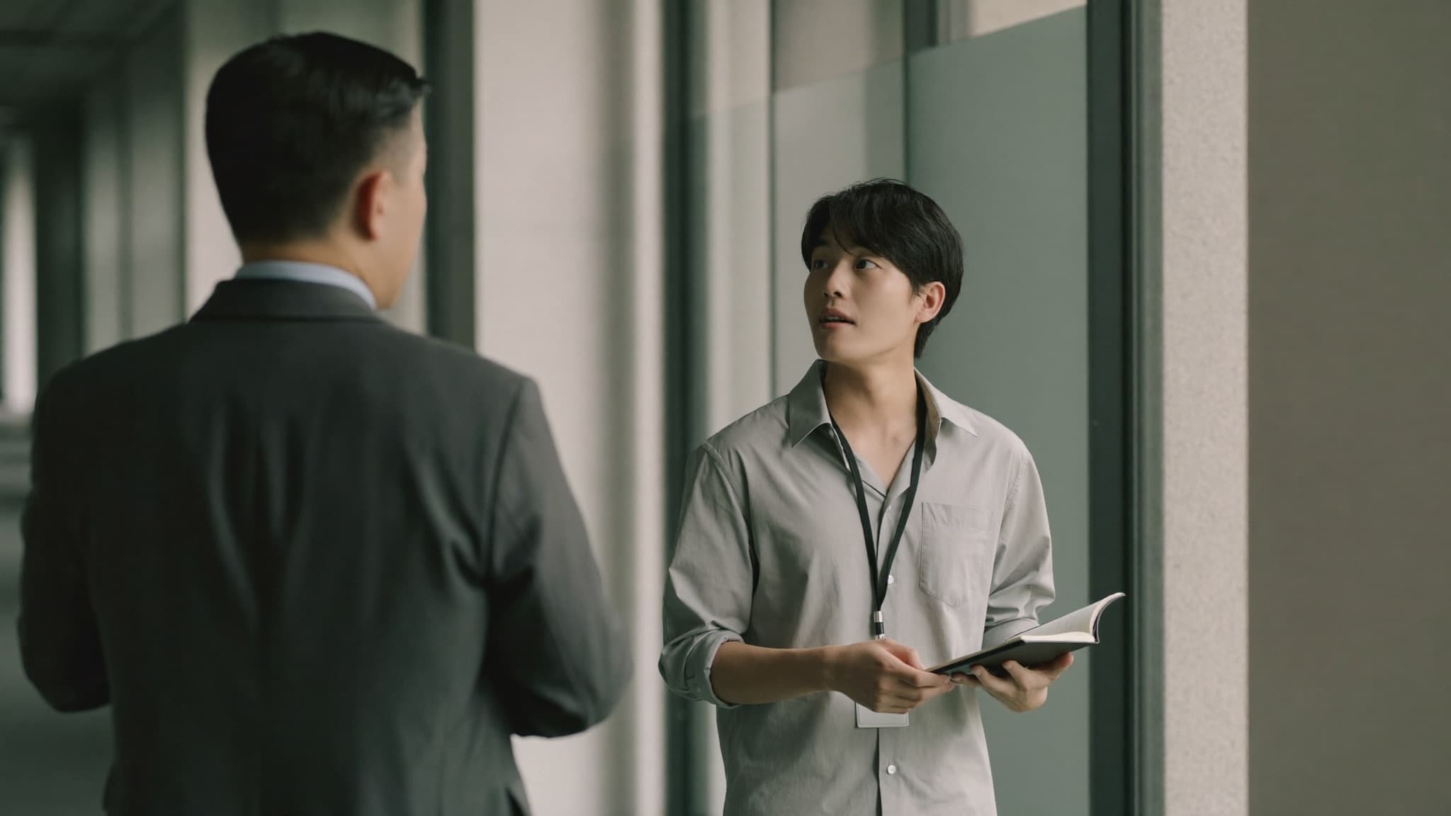 Two colleagues talking in an office corridor: an older one in a suit with their back to the camera, a younger one holding a notebook and a lanyard, looking up as they answer.