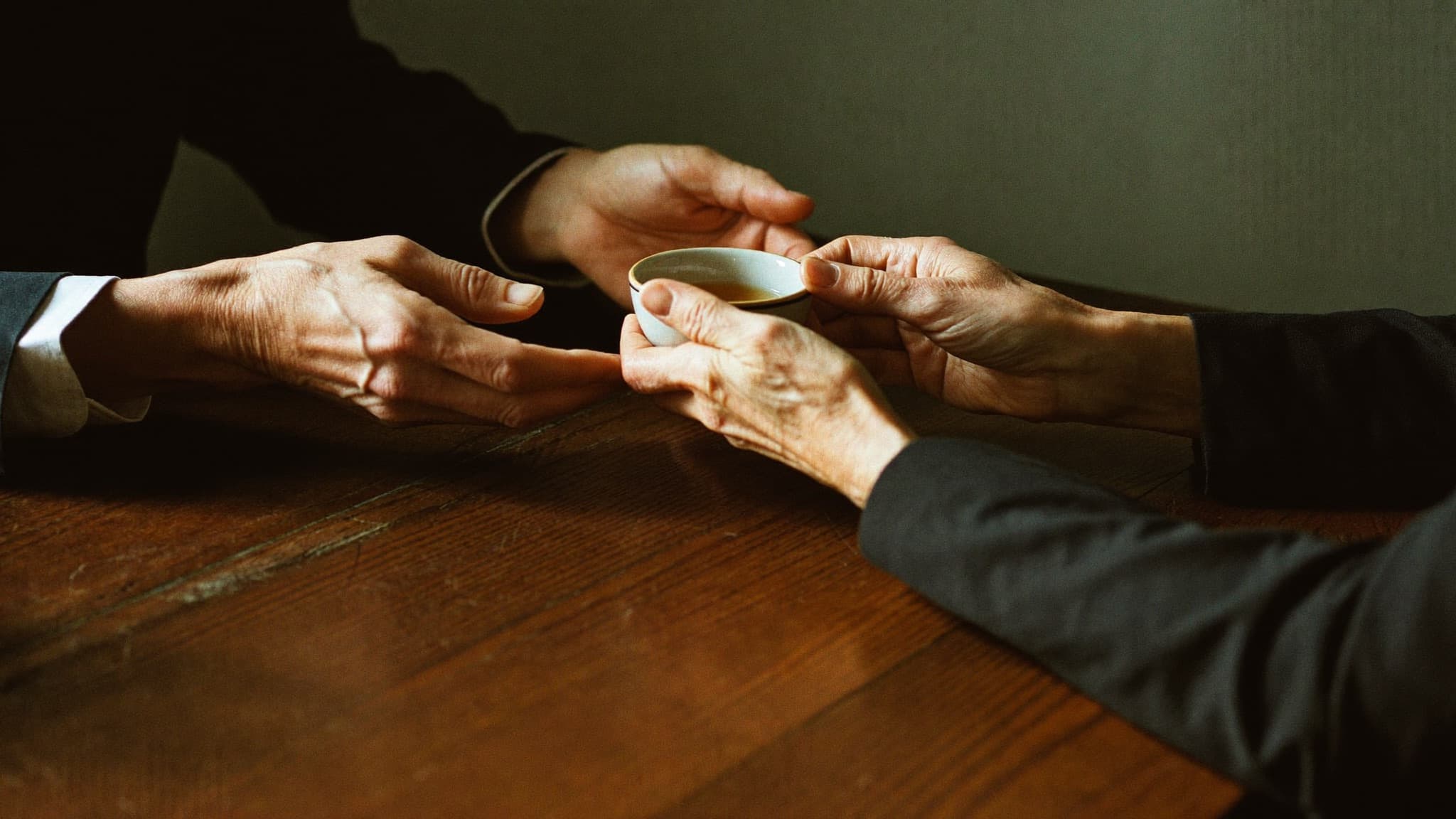 Close view across a wooden table of a small cup of tea being offered and received, each person using both hands.